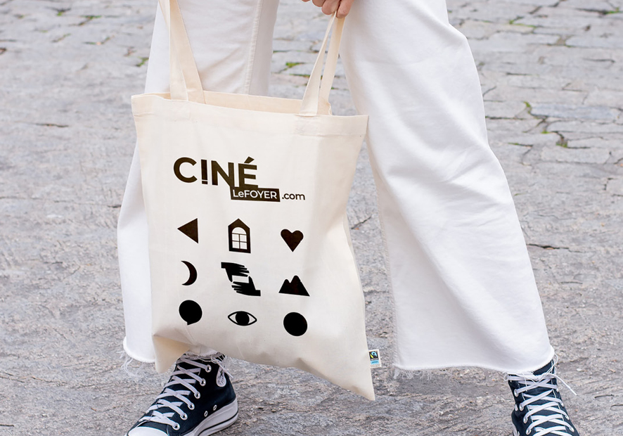 femme en pantalon blanc et basket converse noires sur route pavee qui porte un tote bag a la main avec les pictos et le nouveau logo du cinema le foyer de bourg argental pilat 42